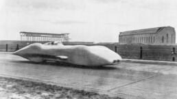 Rudolf Caracciola stellt am 28. Januar 1938 auf der Autobahn Frankfurt–Darmstadt mit 432,7 km/h im Mercedes-Benz V12-Rekordwagen W 125 einen Geschwindigkeitsweltrekord für öffentliche Straßen auf. Foto während der Rekordfahrt, im Hintergrund die Zeppelinhalle bei Frankfurt am Main. (Fotosignatur der Mercedes-Benz Classic Archive: R5682)