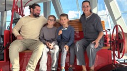 Bettina Haussmann überreicht dem 14-millionsten Besucher des Mercedes-Benz Museums, Sebastian N. aus Aspach, mit Sohn Benedikt und Freund Flynn ein Geschenk – Erinnerungsfoto vor historischen Feuerwehrfahrzeugen.