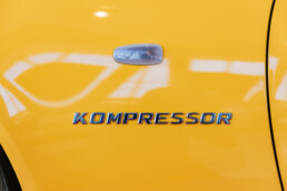 Mercedes-Benz SLK 230 Kompressor der Baureihe R 170 in der Lackierung yellowstone (Mercedes-Benz Farbcode 685) aus dem Jahr 1996. Detailaufnahme des „KOMPRESSOR“-Schriftzugs auf dem linken Kotflügel in der Sonderausstellung „Youngtimer“ des Mercedes-Benz Museums, 2025. Foto: Thomas Niedermüller. (Fotosignatur der Archive von Mercedes-Benz Classic: D854695)